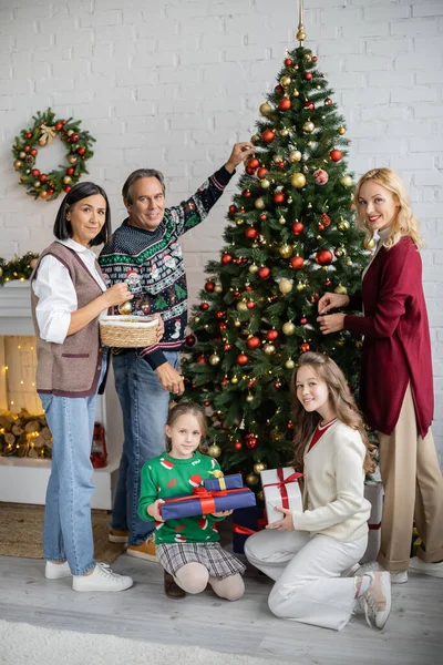 Glad Multietnisk Familj Tittar Kameran Samtidigt Dekorera Julgran Vardagsrummet — Stockfoto