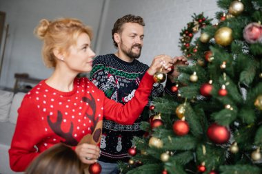 Ailesinin yanında süveter giyen, Noel ağacı süsleyen gülümseyen bir adam. 