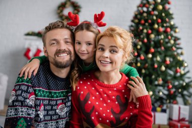 Gülümseyen bir aile Noel süveteri içinde kucaklaşıyor ve evde kameraya bakıyor. 