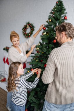 Gülümseyen kız babamın yanında Noel ağacı süslüyor ve annemi evde bulanıklaştırıyor. 
