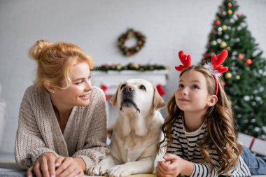 Gülümseyen anne, evde Labrador yakınlarında Noel bandıyla kızına bakıyor. 