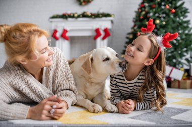 Gülümseyen anne kızına bakıyor. Evde saç bandı ve labrador takıyor. 