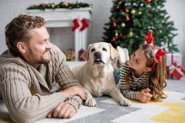 Gülümseyen baba, evde Labrador yakınlarında Noel bandıyla kızına bakıyor. 