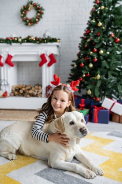 Neşeli kız Labrador 'u okşuyor ve oturma odasındaki Noel ağacının yanındaki kameraya bakıyor. 