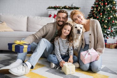 Evde Labrador yakınlarında neşeli bir aile Noel hediyeleri tutuyor. 