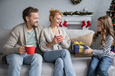 Gülümseyen çocuk elinde noel hediyesiyle ailesinin yanında evdeki kanepede elinde fincanlarla oturuyor. 