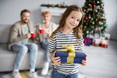 Neşeli kız elinde noel hediyesiyle evde bulanık anne ve babasının yakınındaki kameraya bakıyor. 