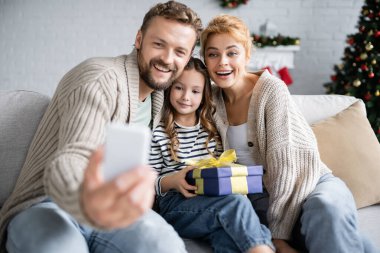 Gülümseyen adam akıllı telefondan evinde Noel hediyesiyle karısının ve kızının yanında selfie çekiyor. 