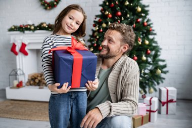 Baba, oturma odasında Noel hediyesiyle mutlu kızına bakıyor.