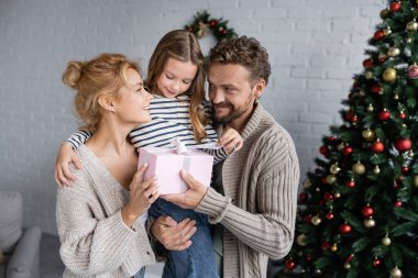 Gülümseyen adam hediyesini tutuyor ve karısının yanında kızı ve evde Noel ağacı. 