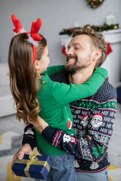 Smiling man in sweater hugging daughter and holding christmas gift at home