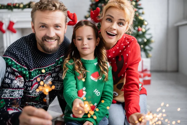 Alegres Padres Sosteniendo Destellos Borrosas Cerca Hija Durante Navidad Casa —  Fotos de Stock