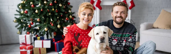 笑顔カップルで暖かいクリスマスのセーターでカメラを見て近くのラブラドール自宅 バナー — ストック写真