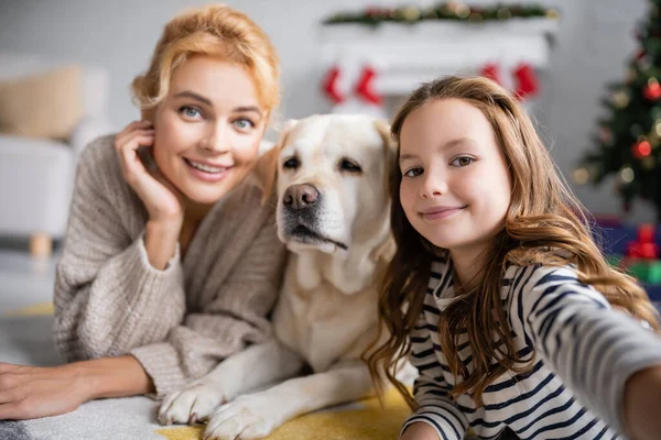 Smiling girl looking at camera near mom and labrador on floor at home