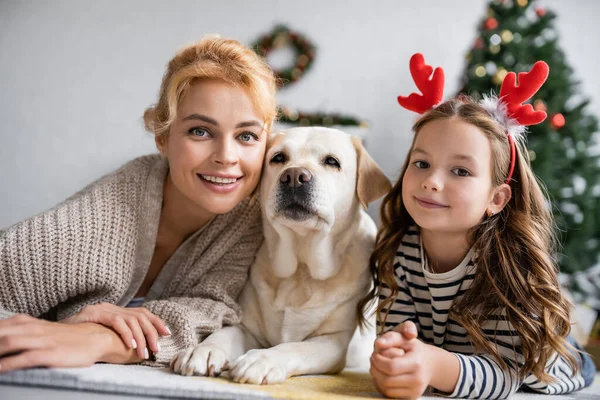 Positive mom and daughter with christmas headband looking at camera near labrador on floor at home