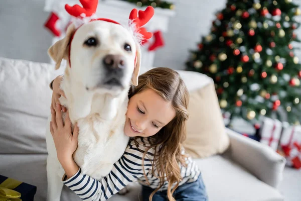 Wesoła Dziewczyna Przytula Labradora Świąteczną Opaską Kanapie Salonie — Zdjęcie stockowe
