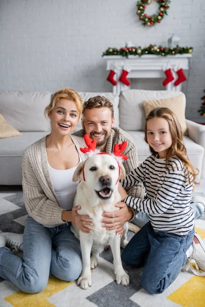 Happy family looking at camera near blurred labrador with christmas headband in living room