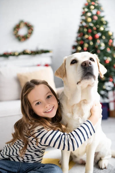 Chica Sonriendo Cámara Mientras Abraza Labrador Cerca Borrosa Árbol Navidad — Foto de Stock