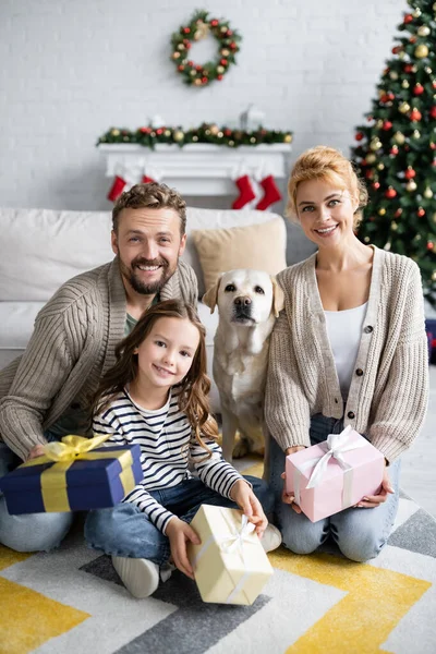 Positiva Familj Hålla Gåvor Och Titta Kameran Nära Labrador Hemma — Stockfoto