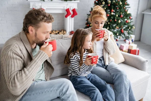 Woman looking at daughter with cup of cacao near husband on couch during new year at home