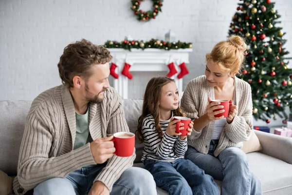 Smiling parents looking at daughter with cacao on couch during new year