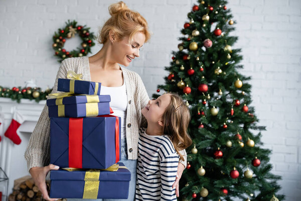 Вид сбоку улыбающейся женщины, держащей подарки рядом с дочерью и елкой дома 
