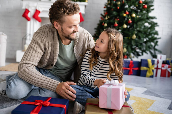 Улыбающийся мужчина обнимает дочь возле рождественских подарков дома 