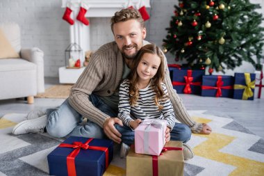 Gülümseyen adam ve kız hediyelerin yakınındaki kameraya bakıyor ve evde bulanık Noel dekorasyonu yapıyor. 