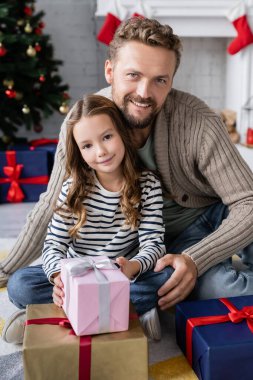 Gülümseyen adam ve kızı evdeki Noel hediyelerinin yakınında kameraya bakıyor. 