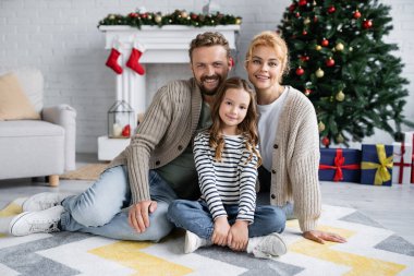 Mutlu ebeveyn ve kız evde bulanık Noel ağacının yanında kamerada gülümsüyor. 