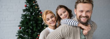 Sıcak hırka giymiş olumlu aile evdeki Noel ağacının yakınındaki kameraya bakıyor, afiş. 