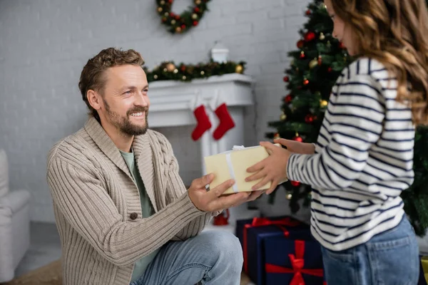 Happy man in knitted cardigan giving gift box to blurred daughter during christmas at home