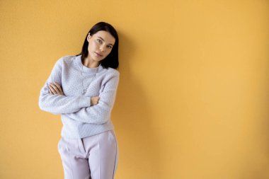 Güzel esmer model, rahat örgü süveter, çapraz kollarla poz veriyor ve sarı arka plandaki kameraya bakıyor.