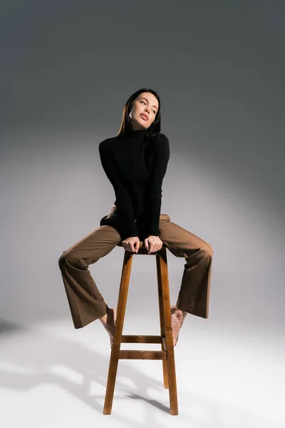 穿着棕色裤子和黑色毛衣的性感赤脚女人坐在灰色背景的高凳子上 — 图库照片