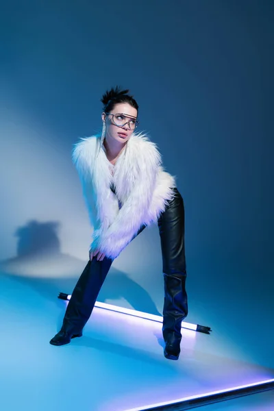 brunette model in black leather pants and white faux fur jacket looking away near fluorescent lamp on blue background