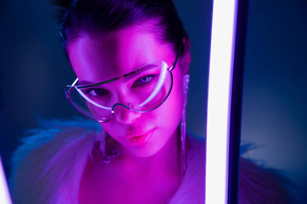 Модная женщина в солнцезащитных очках смотрит в камеру рядом с лампой на темно-синем фоне 