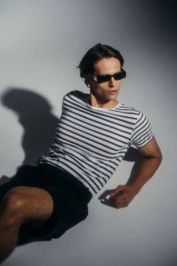 Şortlu ve güneş gözlüklü esmer adamın, gri arka planda, gölgesi olan yüksek açılı görüntüsü. 