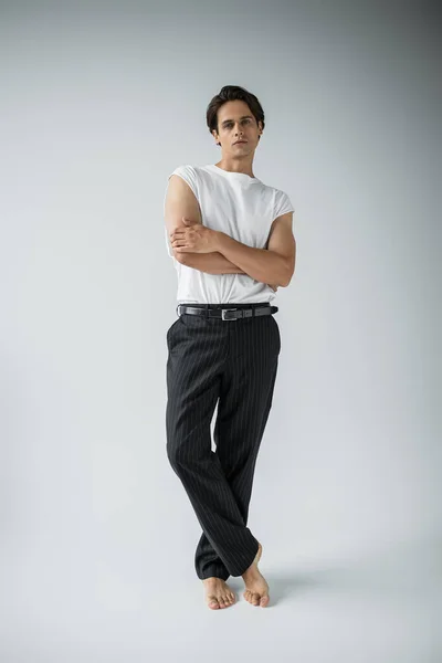 full length of barefoot man in striped trousers and white t-shirt posing with crossed arms on grey