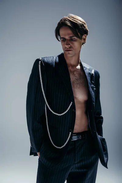 stylish man with hairy chest and suit with pearl chain posing with hands in pockets on dark grey