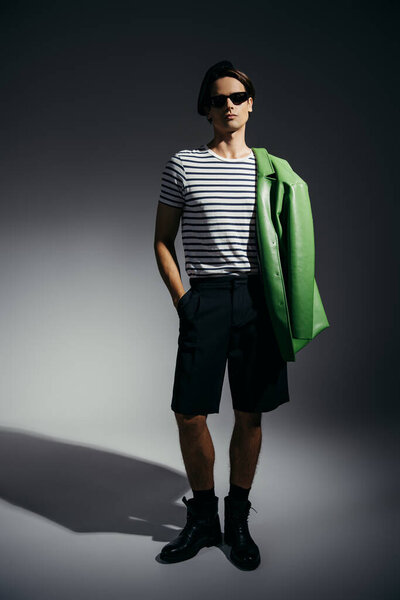 Full length of stylish man in sunglasses and shorts holding leather jacket on shoulder on grey background with lighting 