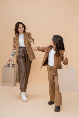 Alışveriş çantalı moda kız bej arka planda neşeli ve şık annesiyle el ele tutuşuyor.