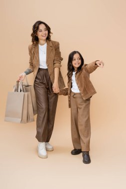 Gülümseyen kız moda annesiyle el ele tutuşuyor alışveriş torbalarıyla ve bej rengi parmağıyla işaret ediyor. 