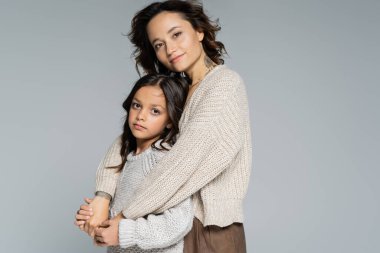 Dövmeli esmer kadın, esmer kıza sarılıyor ve gri renkli kameraya gülümsüyor.