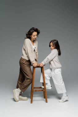 Gri üzerine ahşap tabureye yakın poz veren modaya uygun sonbahar kıyafetli bir kadın ve kız. 