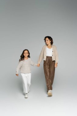 Gri arka planda el ele tutuşurken anne ve kızının tam boy örgü ve pantolon giymesi.