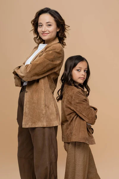 mom and daughter in trendy autumn outfit posing back to back with crossed arms and looking at camera isolated on beige