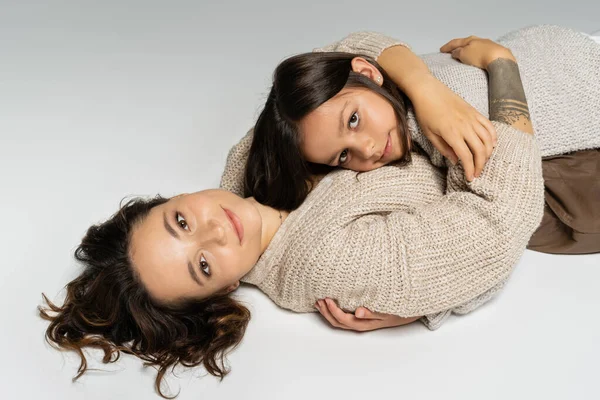 brunette mom and daughter in cozy knitwear smiling at camera and hugging on grey background