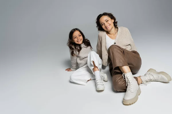 トレンディで暖かい服を着た女の子と母親が灰色の背景のカメラに座って笑っています — ストック写真
