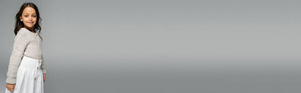 веселый ребенок в вязаном свитере и белой юбке позирует изолированно на сером, баннер