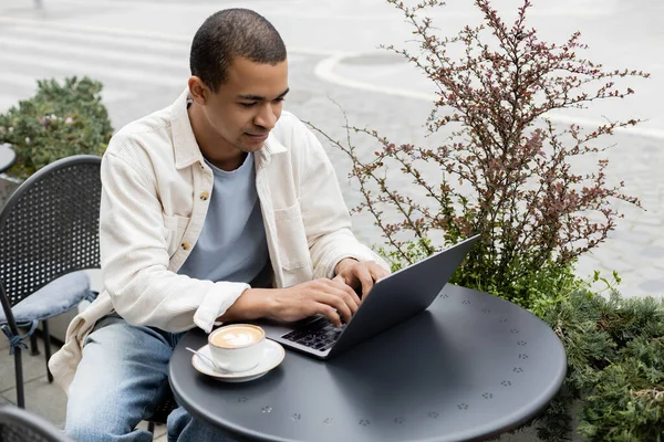 Hombre afroamericano usando portátil cerca de taza de café con leche con espuma en la mesa en la terraza de la cafetería - foto de stock